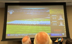 People gathered around a big screen TV with an image of a sunny baseball field on it, with a big American flag being paraded around on the grass